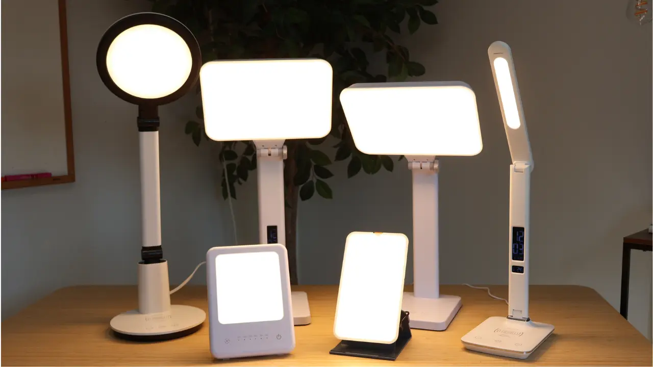 Six different modern light therapy lamps of various shapes and sizes are displayed on a wooden table. Each lamp emits a bright white light, and a leafy plant is visible in the background.