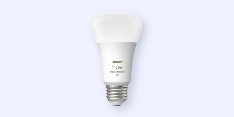 A Philips Hue LED smart light bulb with a white base and silver screw fitting, labeled hue white and color 100, set against a light blue background.