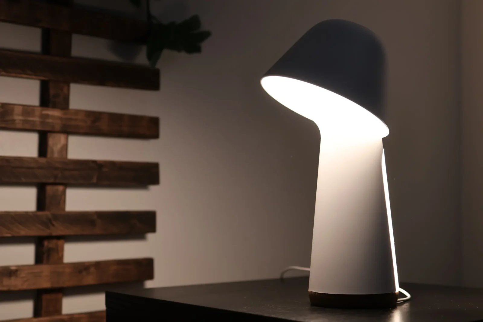 A modern, sculptural white table lamp with a tilted shade emits a soft light, sitting on a dark surface with a wooden slatted wall and plant leaves in the background.