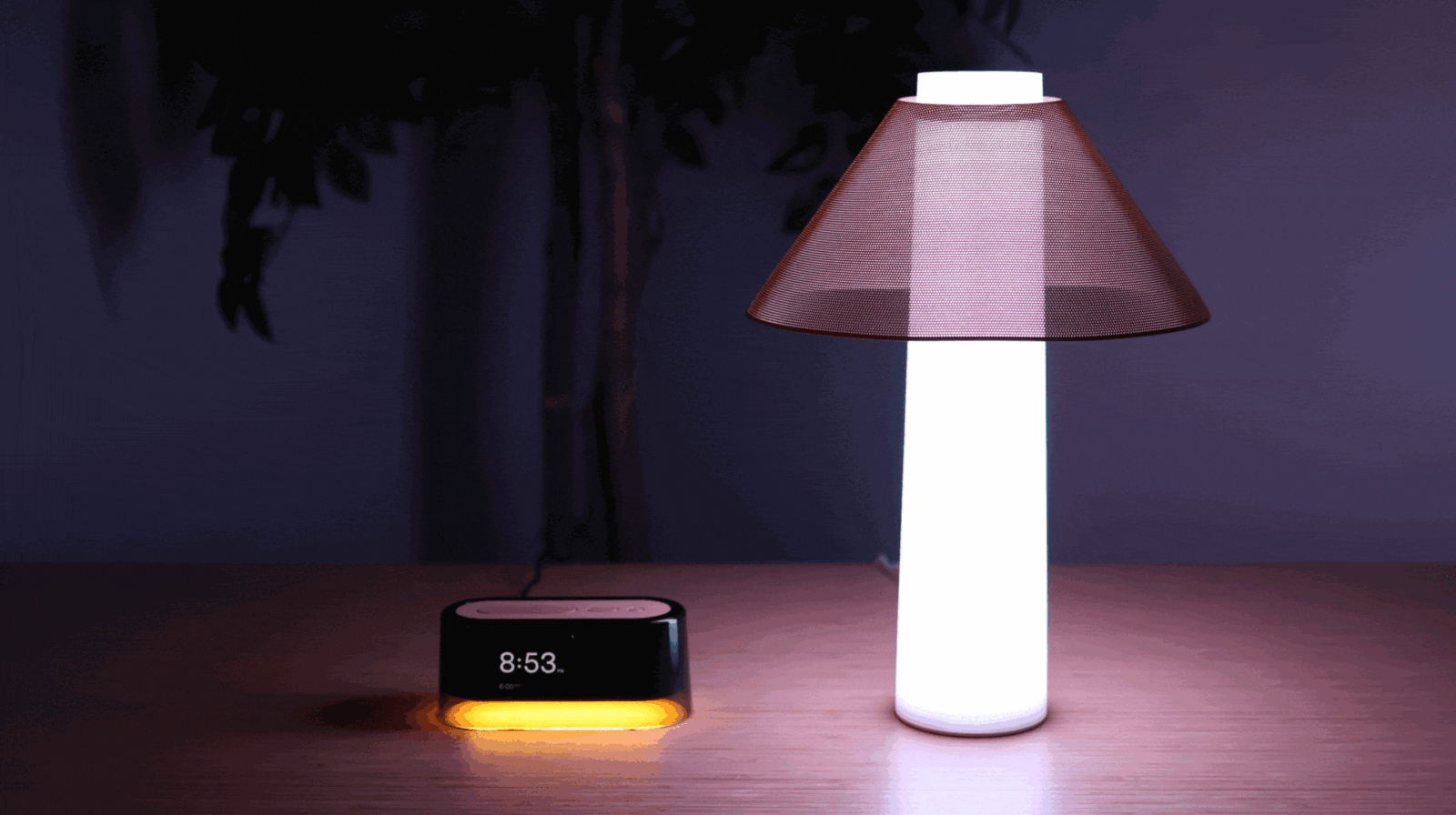 A modern digital alarm clock displaying 8:53 and glowing orange sits on a wooden table next to a tall, cylindrical lamp with a translucent, cone-shaped shade. The lamp emits a bright white light.