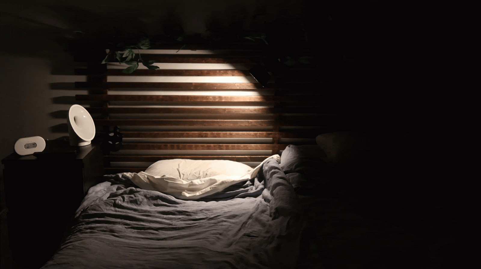 A dimly lit bedroom with a wooden slat headboard, a single pillow lit by a focused light, and unmade bedding. A nightstand with a small round lamp and a digital clock is next to the bed.