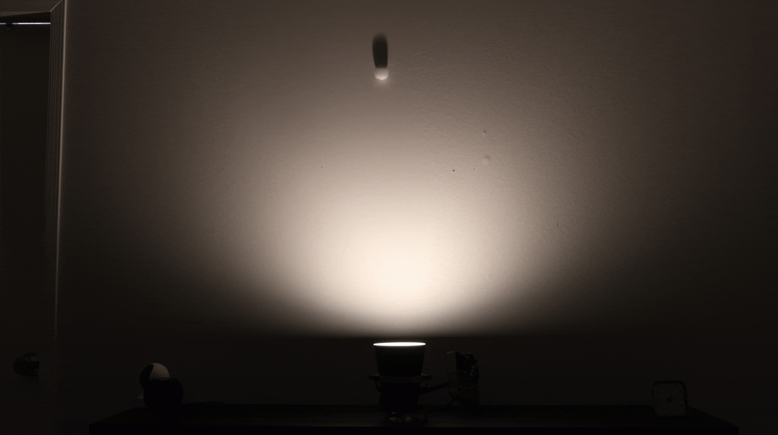 A spotlight on a dark table projects a focused beam of light onto a wall, illuminating the wall in a circular pattern with a shadow of a small object above the light.