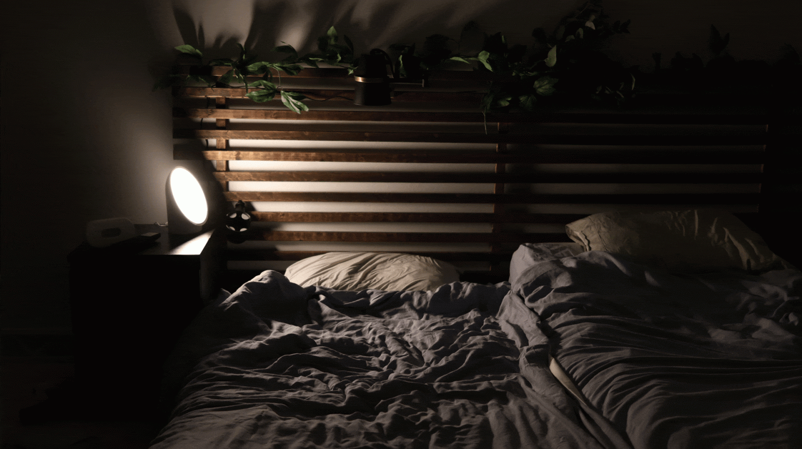 A dimly lit bedroom with a wooden slatted headboard, two pillows, unmade bedding, a nightstand, a glowing round lamp, and green plants on the headboard shelf.