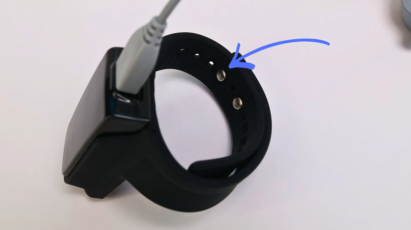 A smart fitness watch with a black wristband is being charged. A blue arrow points to metal contact points on the inside of the band.