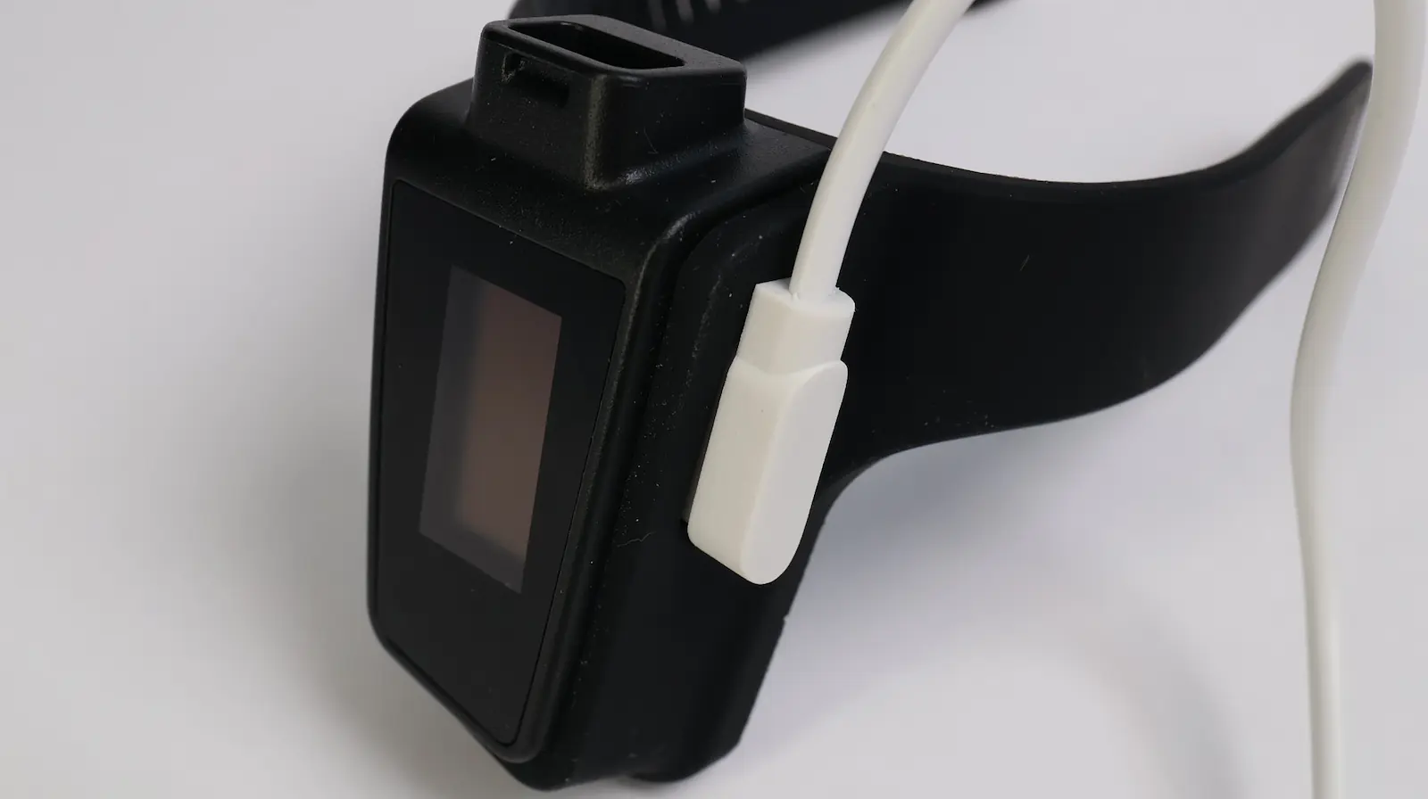 A black smartwatch is shown on a white surface, with a white charging cable connected to its side. The screen of the watch is off or inactive.