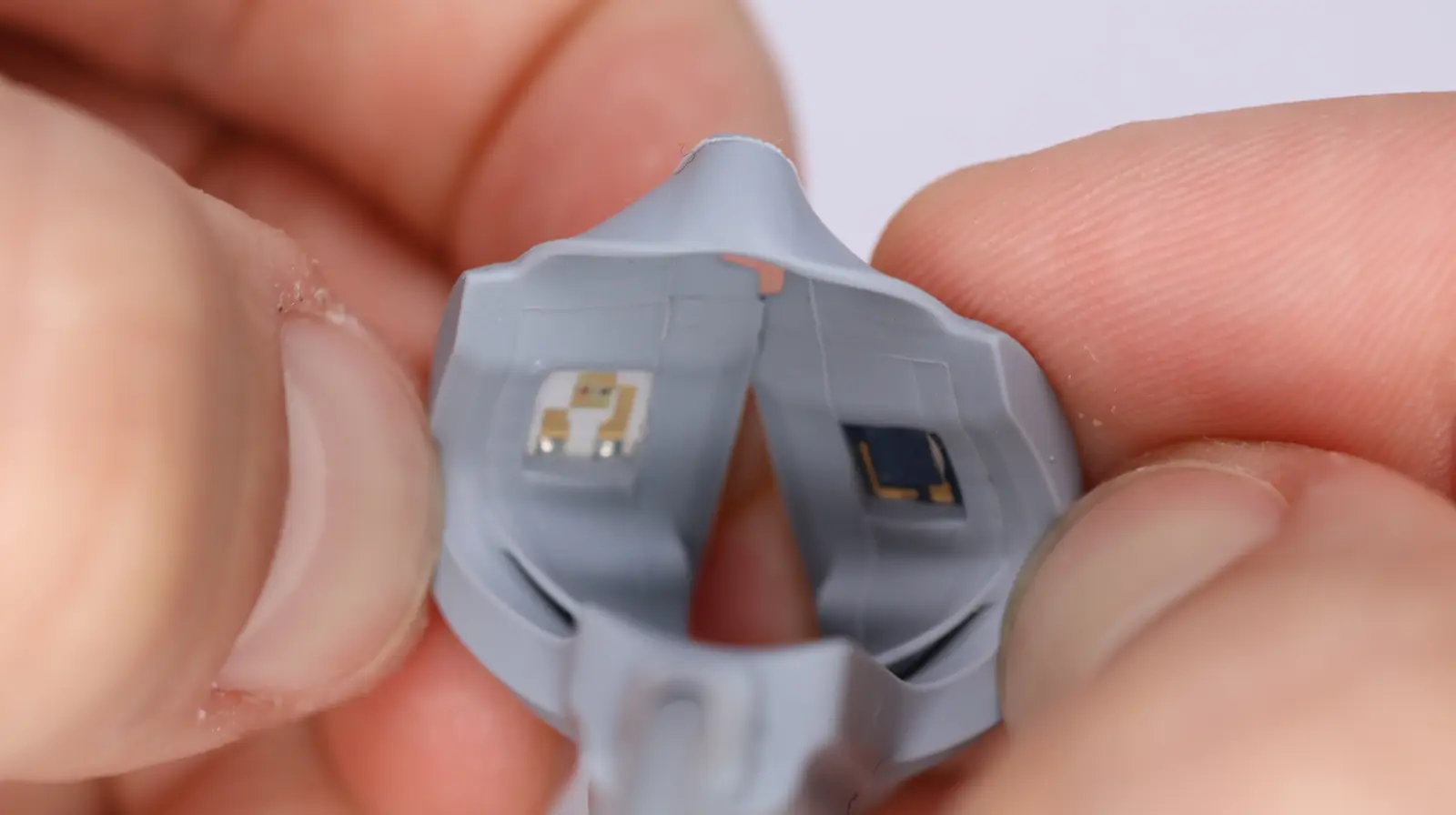 Close-up of two hands holding a small gray object with two tiny electronic components or sensors attached inside. The components are square-shaped and positioned side by side.
