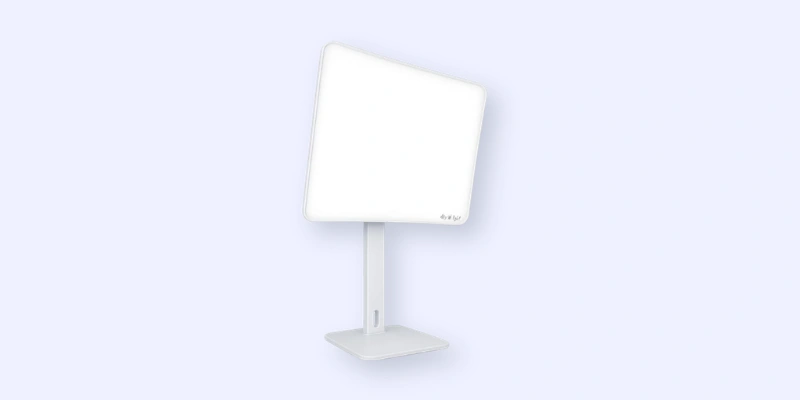 A rectangular white light therapy lamp with an angled face, mounted on a slim, adjustable stand and base, against a light blue background.