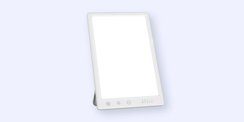 A rectangular white light therapy lamp with a touchscreen panel at the bottom featuring power, brightness, and timer controls, standing at an angle against a light background.