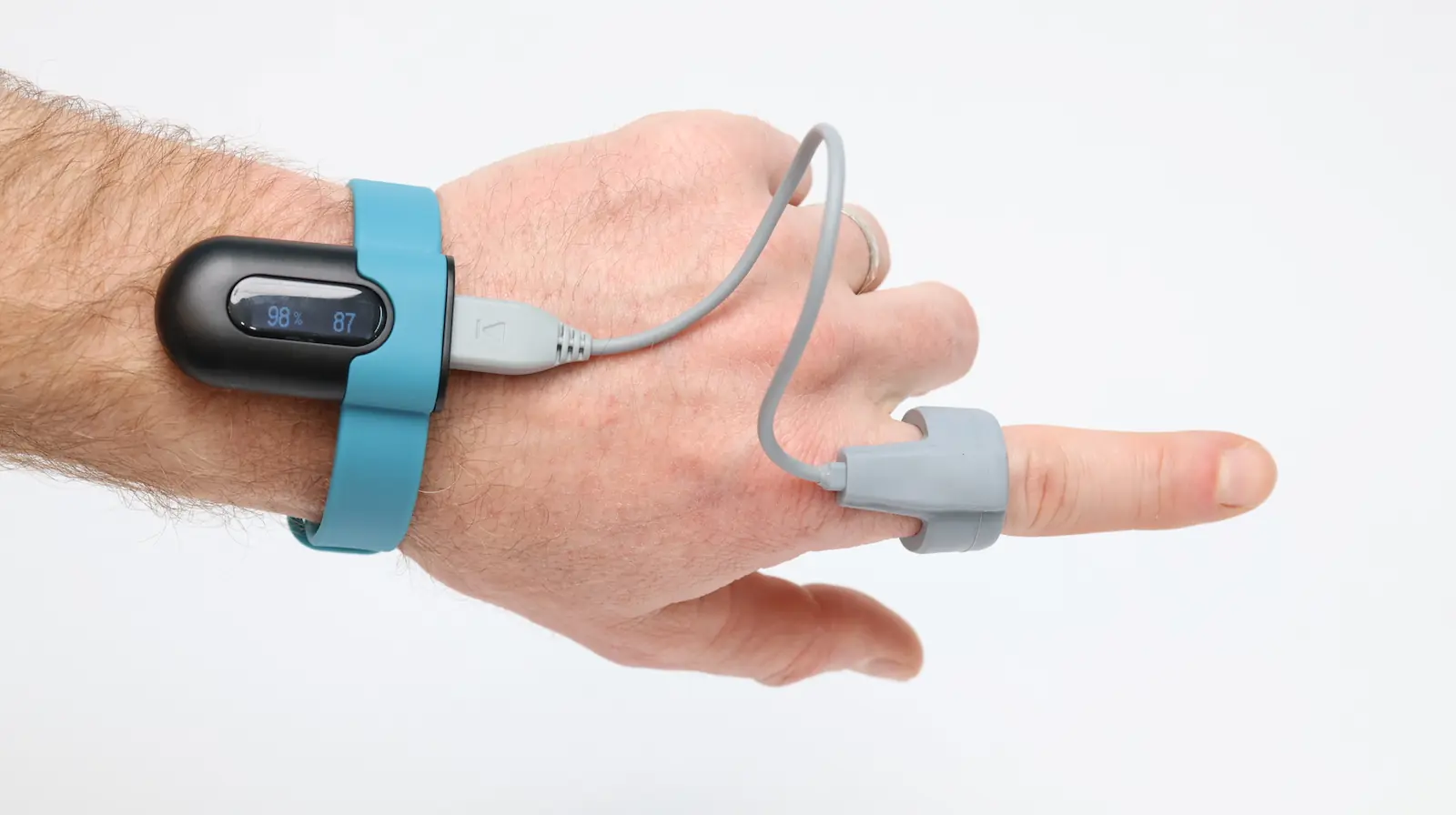 A hand wearing a blue wrist device connected by a cable to a gray sensor clipped onto the index finger, displaying readings on a small screen against a plain white background.