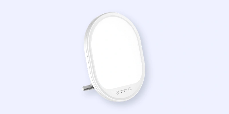 A small, oval-shaped light therapy lamp with a stand and touch controls at the bottom, set against a light blue background.