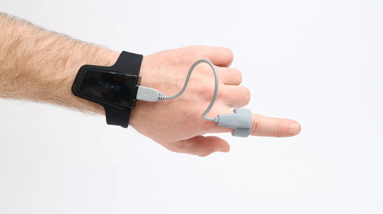 A hand wearing a black wrist device connected by a gray cable to a sensor clipped on the index finger, against a plain white background.