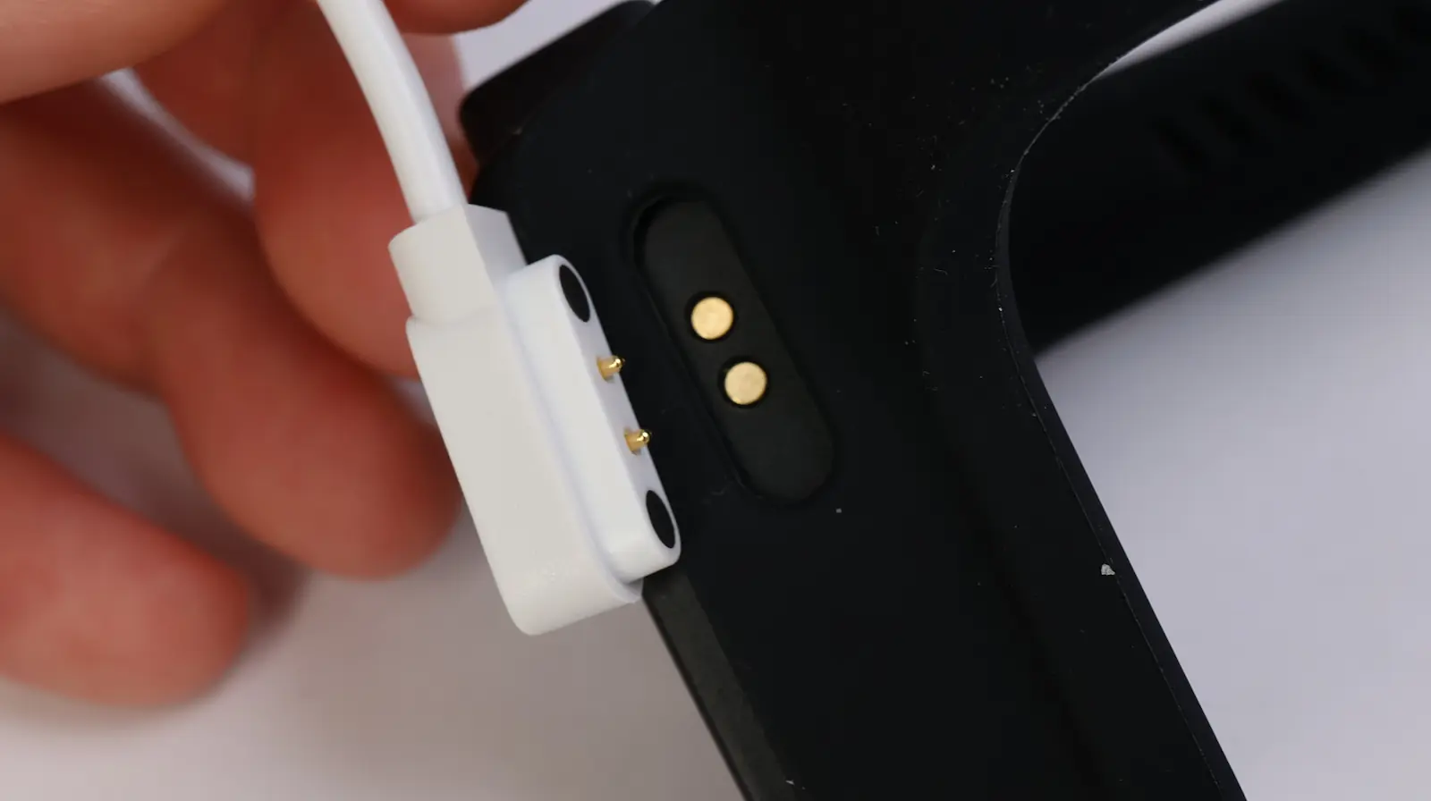 A close-up of a hand holding a white magnetic charger near the charging pins on the back of a black fitness tracker or smartwatch.