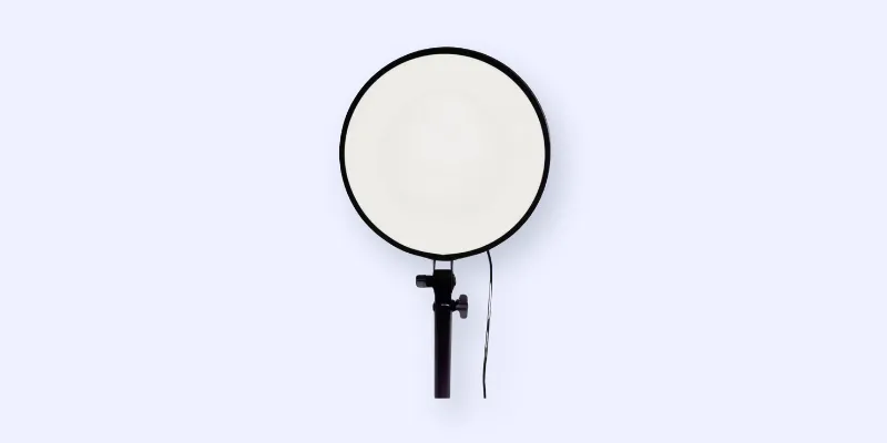 A round, white ring light on a black stand is centered against a plain, light gray background. A black power cord hangs from the light.