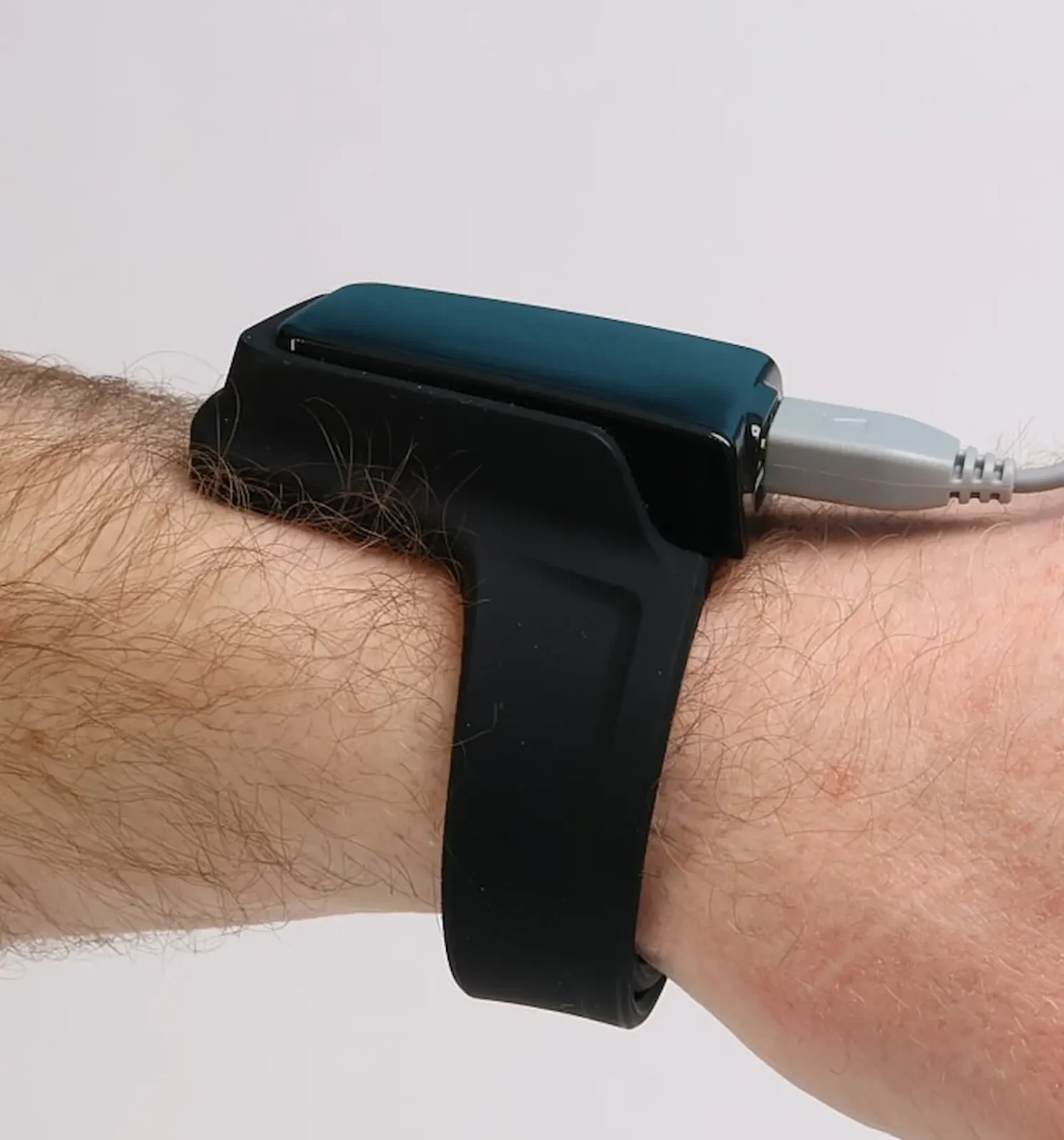 A close-up of a persons wrist wearing a black wristband device with a rectangular top, connected to a gray cable. The background is plain and white.
