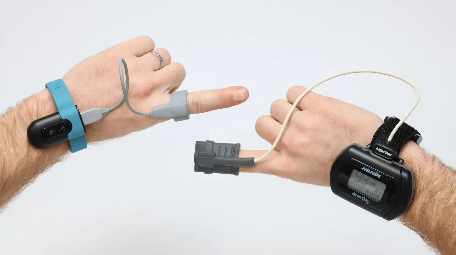 Two hands display different pulse oximeters attached to the index fingers. Each hand wears a wrist device connected by a wire to a sensor clipped on the finger, showing how the devices are used to measure blood oxygen levels.
