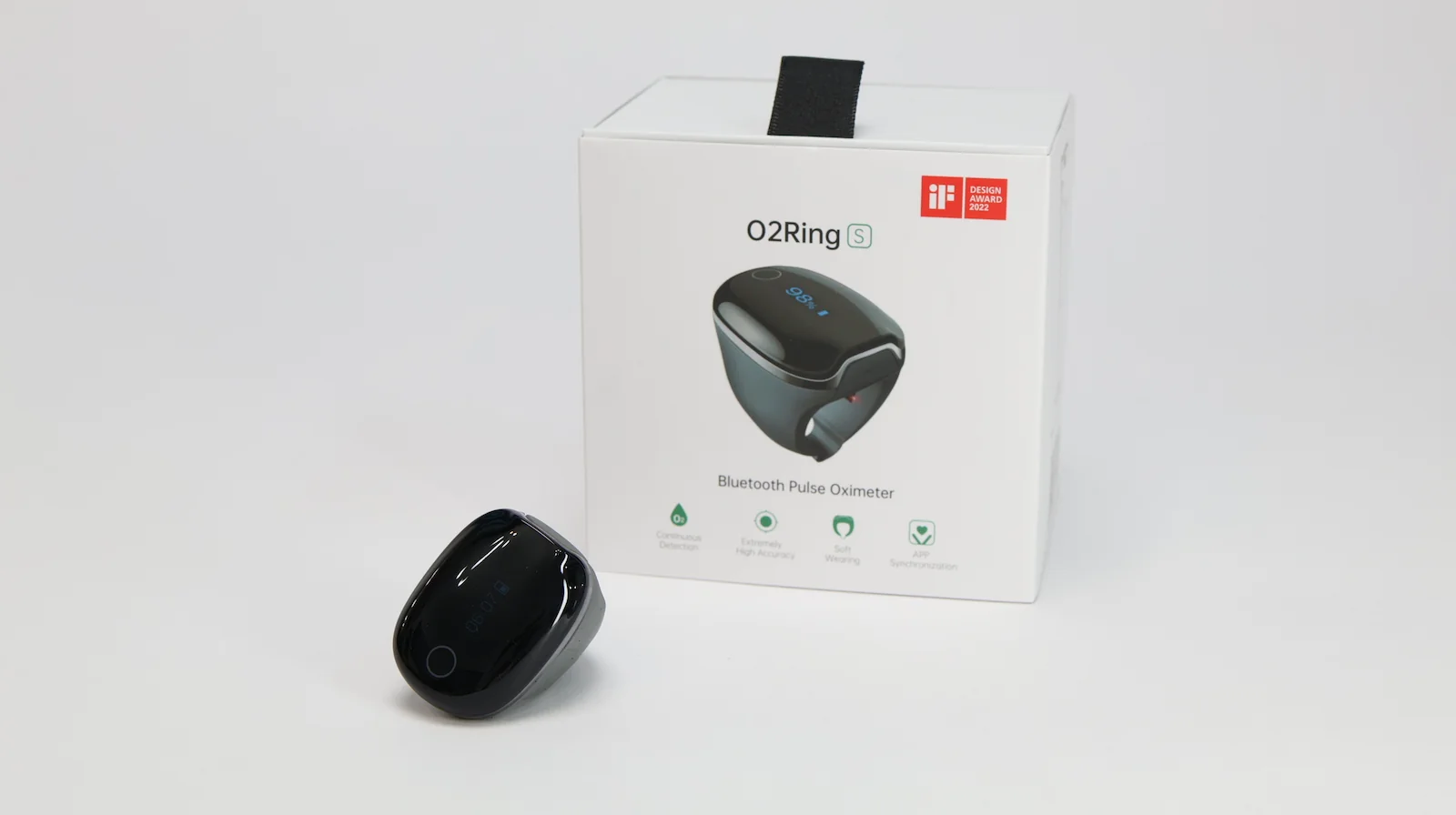 A black O2Ring Bluetooth pulse oximeter is placed in front of its white product box, which features an image of the device and icons indicating key functions on a plain white background.