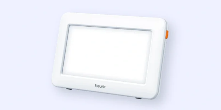 A rectangular white light therapy lamp with a glowing screen, rounded edges, and an orange button on the side, standing upright on small legs. The brand name beurer is visible below the screen.
