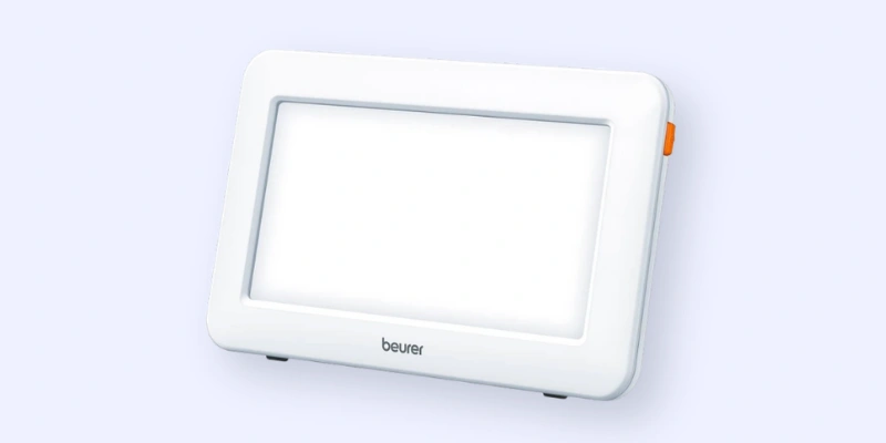 A rectangular white light therapy lamp with a glowing screen, rounded edges, and an orange button on the side, standing upright on small legs. The brand name beurer is visible below the screen.
