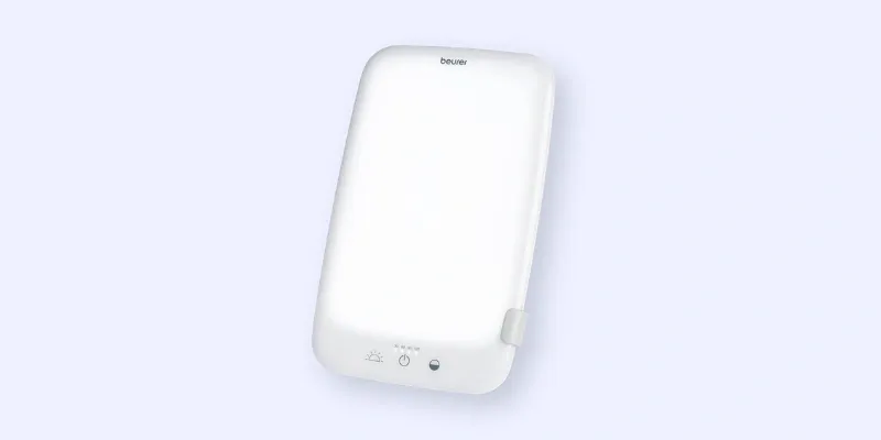 A rectangular white light therapy lamp with a minimalist design, featuring a small button and indicator lights at the bottom front panel, against a light blue background.
