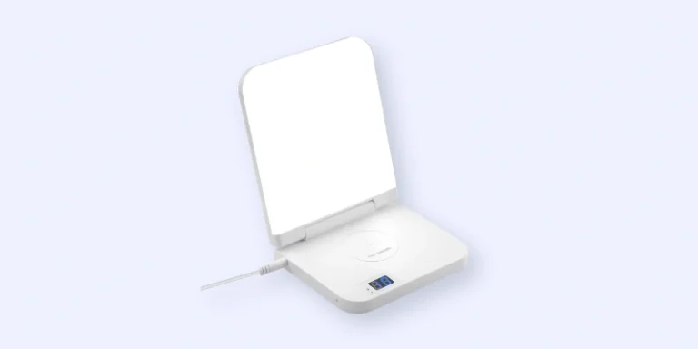 A white, rectangular device with a hinged, illuminated mirror standing upright. The base features a circular touchpad, a digital display, and a charging cable connected on the left side.