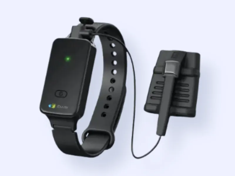 A black wearable device with a wristband, a small rectangular unit with a green indicator light, and a connected clip-on sensor attached by a cable, displayed on a light background.