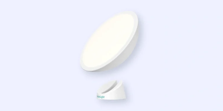 A circular white light therapy lamp with a stand, shown against a light blue background. The lamp is slightly tilted, and the base is separated, displaying the products sleek and modern design.