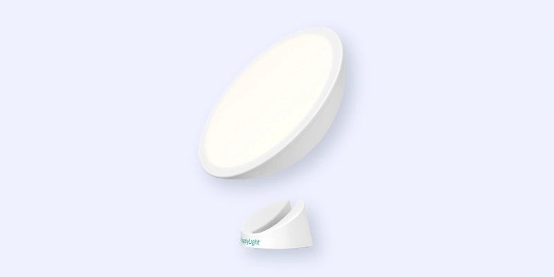 A circular white light therapy lamp with a stand, shown against a light blue background. The lamp is slightly tilted, and the base is separated, displaying the products sleek and modern design.