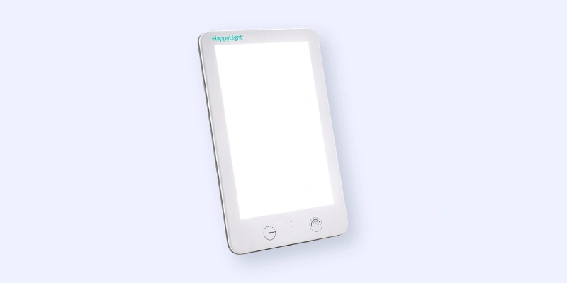 A rectangular white light therapy lamp labeled “HappyLight,” shown angled against a pale background, with touch buttons and a glowing light panel on the front.