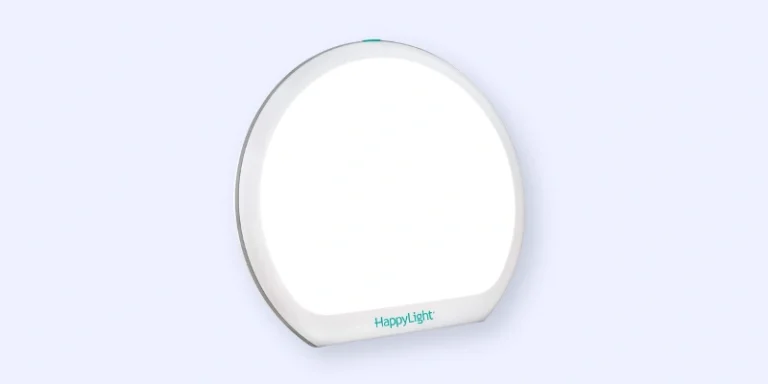 A round, white light therapy lamp with the brand name HappyLight printed on the front, shown turned on against a plain light blue background.