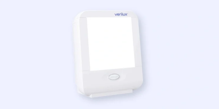 A white rectangular Verilux light therapy lamp with a large illuminated panel and a rounded button below the light, set against a pale background.