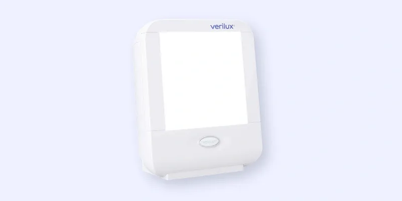 A white rectangular Verilux light therapy lamp with a large illuminated panel and a rounded button below the light, set against a pale background.