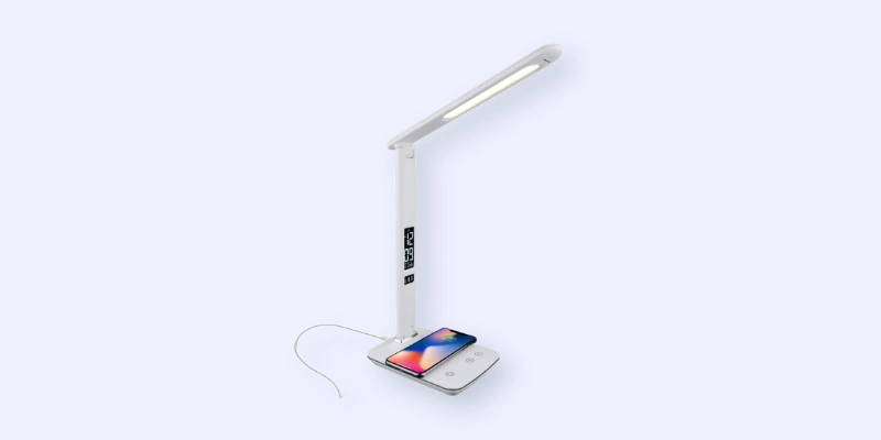 A sleek white desk lamp with an LED light, digital clock display on the stand, and a smartphone charging wirelessly on its square base. A charging cable is connected to the lamp.