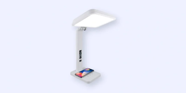 A modern white desk lamp with a rectangular light panel, an adjustable arm, and a base that features wireless phone charging; a smartphone is shown charging on the base.