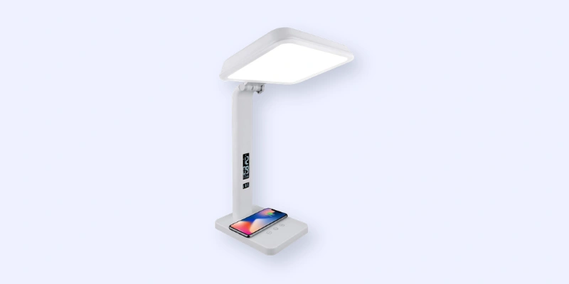 A modern white desk lamp with a rectangular light panel, an adjustable arm, and a base that features wireless phone charging; a smartphone is shown charging on the base.