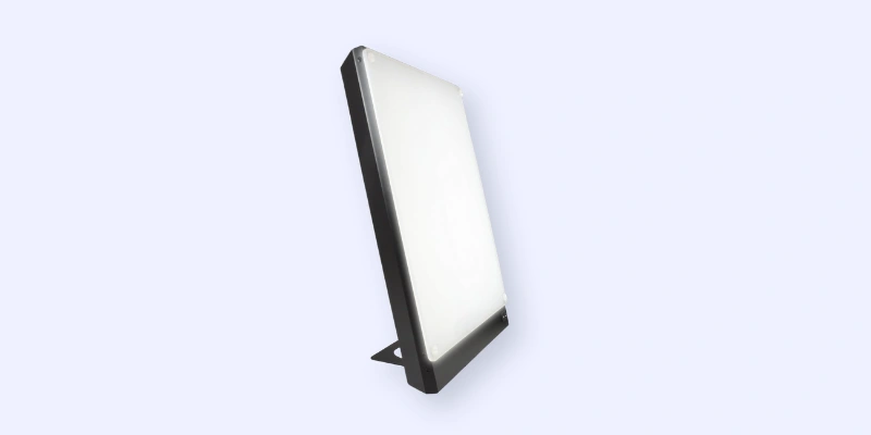 A slim, rectangular LED panel light with a black frame, shown standing upright at an angle on a small built-in stand against a light gray background.