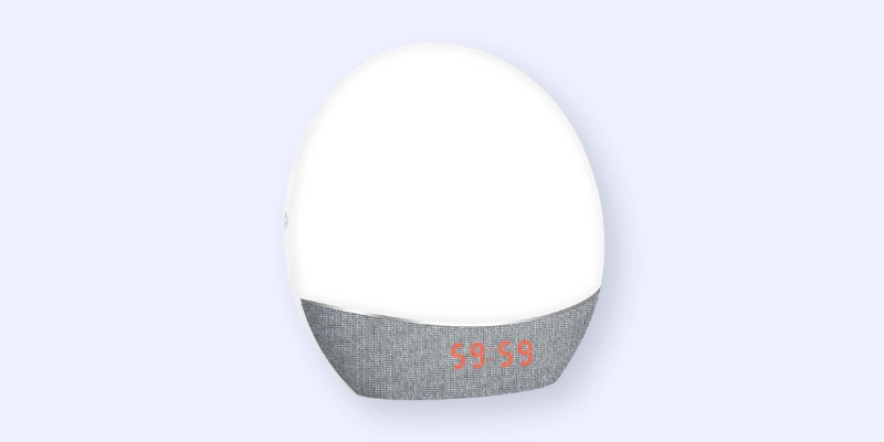 A modern, dome-shaped digital clock with a white upper half and a gray fabric lower half. The time 9:59 is displayed in orange numbers on the bottom section.