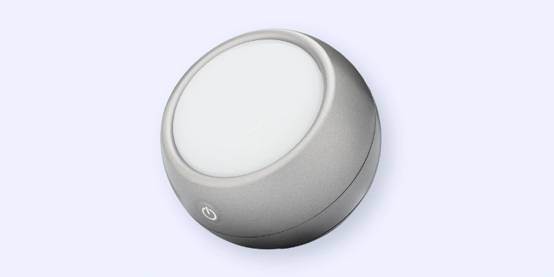 A small, round, gray device with a white circular center and a power button icon on its side, set against a light background. The device appears modern and minimalist in design.