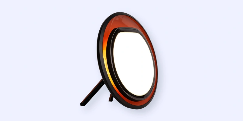 A round, modern tabletop lamp with a bright white light and a circular orange accent, supported by a small stand, set against a pale background.