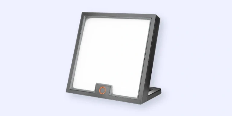 A modern, square-shaped light therapy lamp with a gray frame, a white illuminated screen, and an orange button at the bottom center, set against a light gray background.