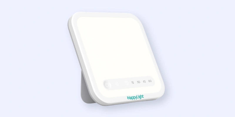 A white rectangular light therapy lamp with rounded edges, standing on a built-in stand. The front panel displays touch controls and a timer. The brand name “HappyLight” is printed at the bottom.