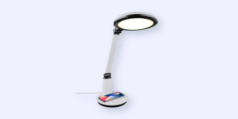 A modern white desk lamp with a round LED light head, an adjustable arm, and a wireless smartphone charging pad at the base. A smartphone is placed on the charging pad.