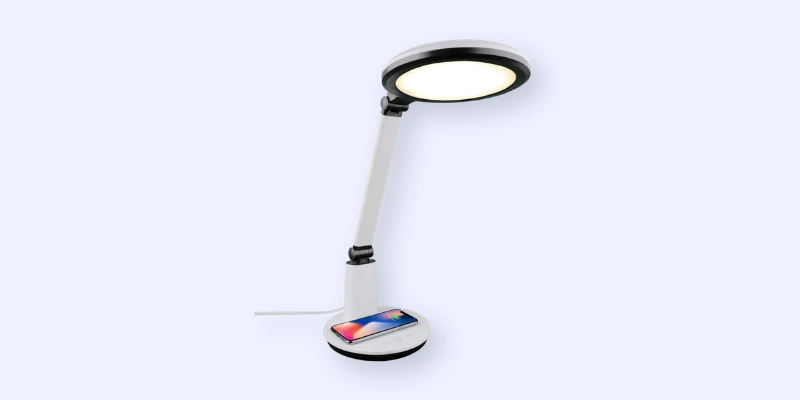 A modern white desk lamp with a round LED light head, an adjustable arm, and a wireless smartphone charging pad at the base. A smartphone is placed on the charging pad.