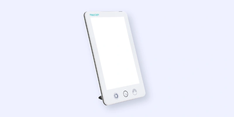 A slim, rectangular light therapy lamp with a white frame and three buttons at the bottom, standing on a built-in stand against a light background. The brand name HappyLight is visible at the top.