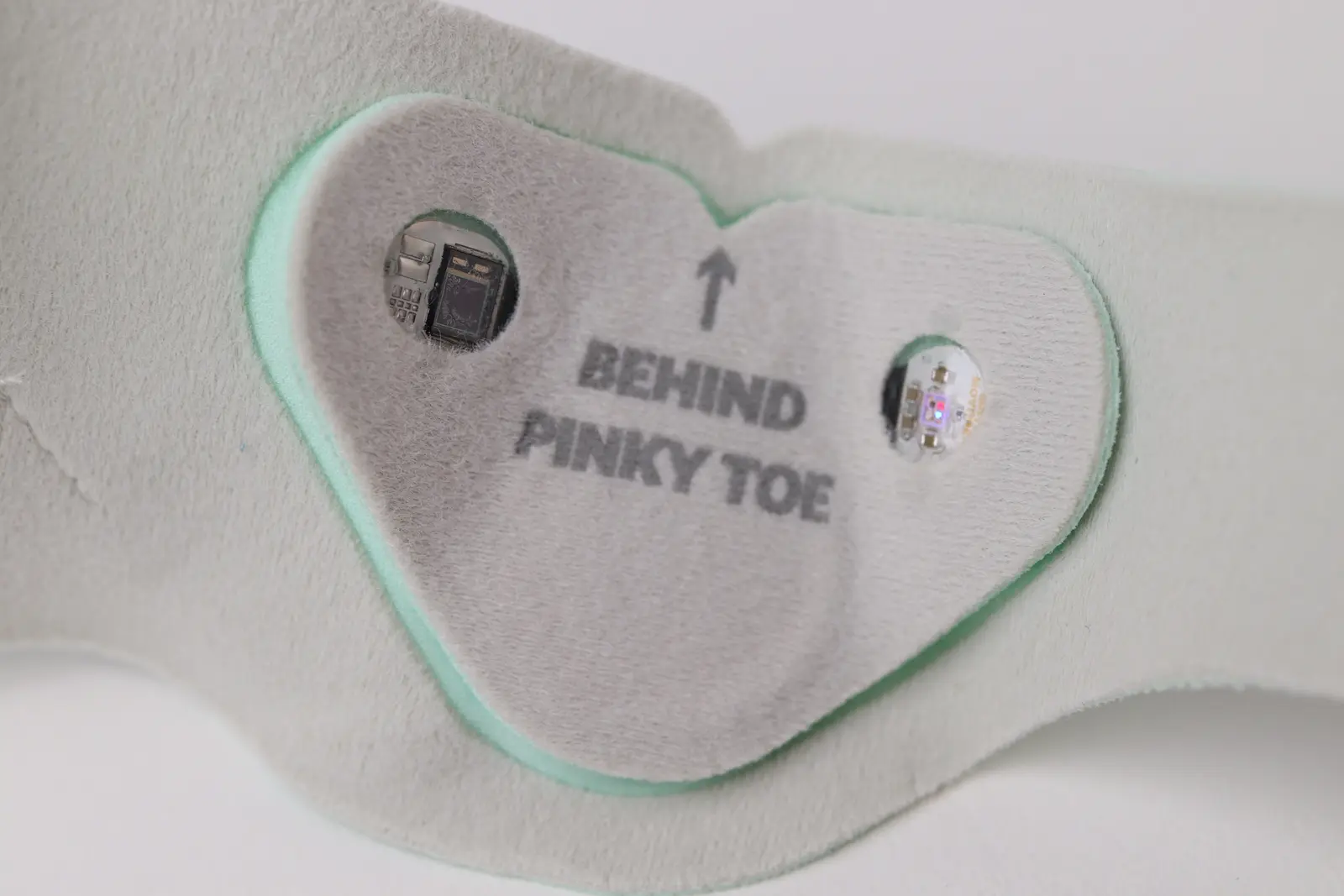 Heart-shaped wearable sensor patch labeled Behind Pinky Toe with embedded microchip and LED components on fabric