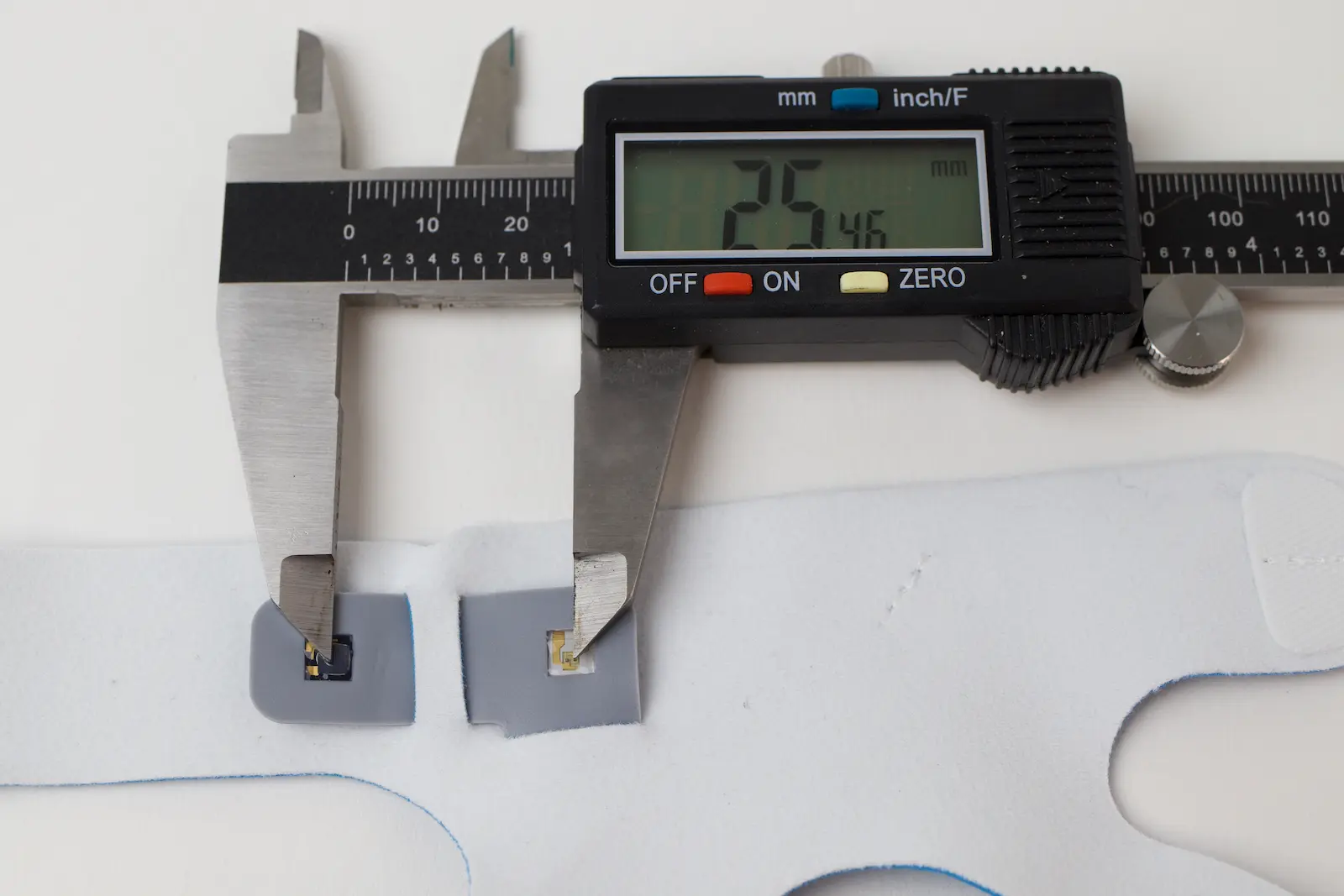 Digital calipers measuring small electronic components on gray pads embedded in a white flexible substrate, reading 25.46mm