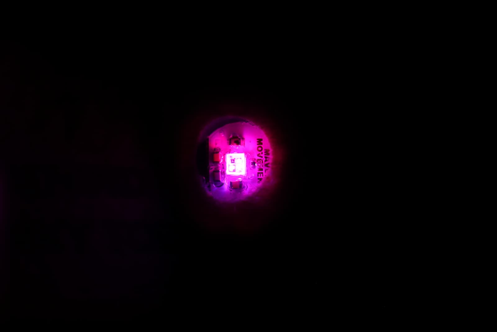 Glowing pink RGB LED on a small circular MAV Movement circuit board illuminated in complete darkness