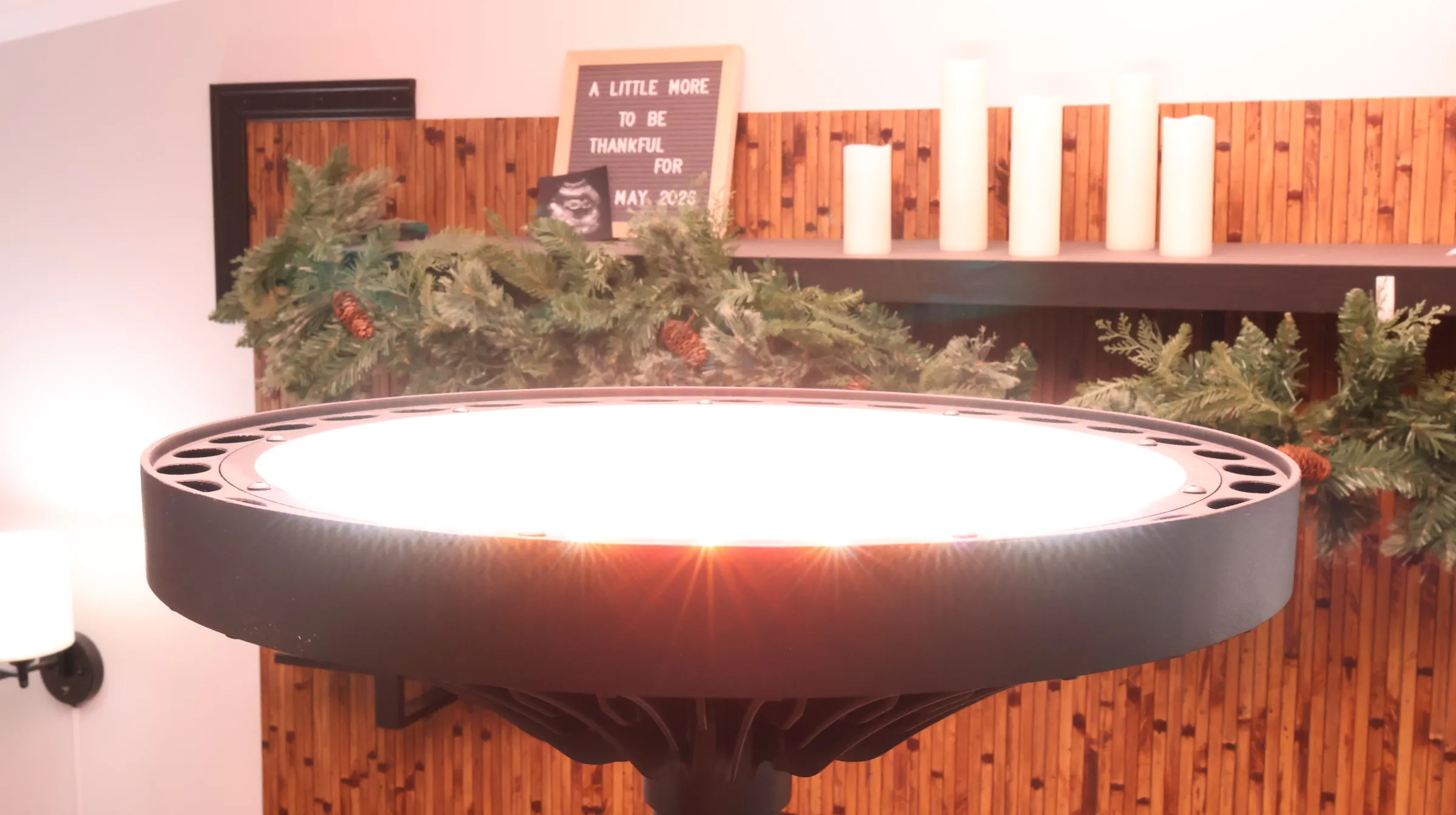 Bright circular LED grow light above a festive shelf with pine garland, white candles, and a pregnancy announcement sign read