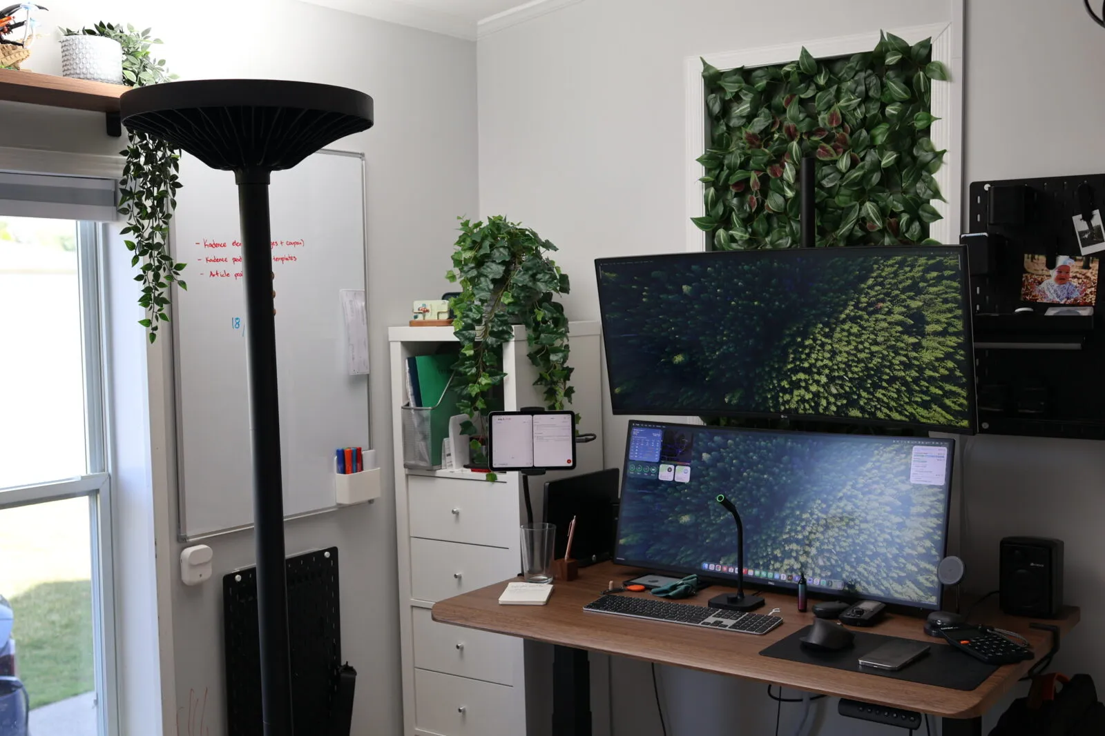 Modern home office with dual monitors, standing desk, whiteboard, and lush green wall plants creating a productive workspace