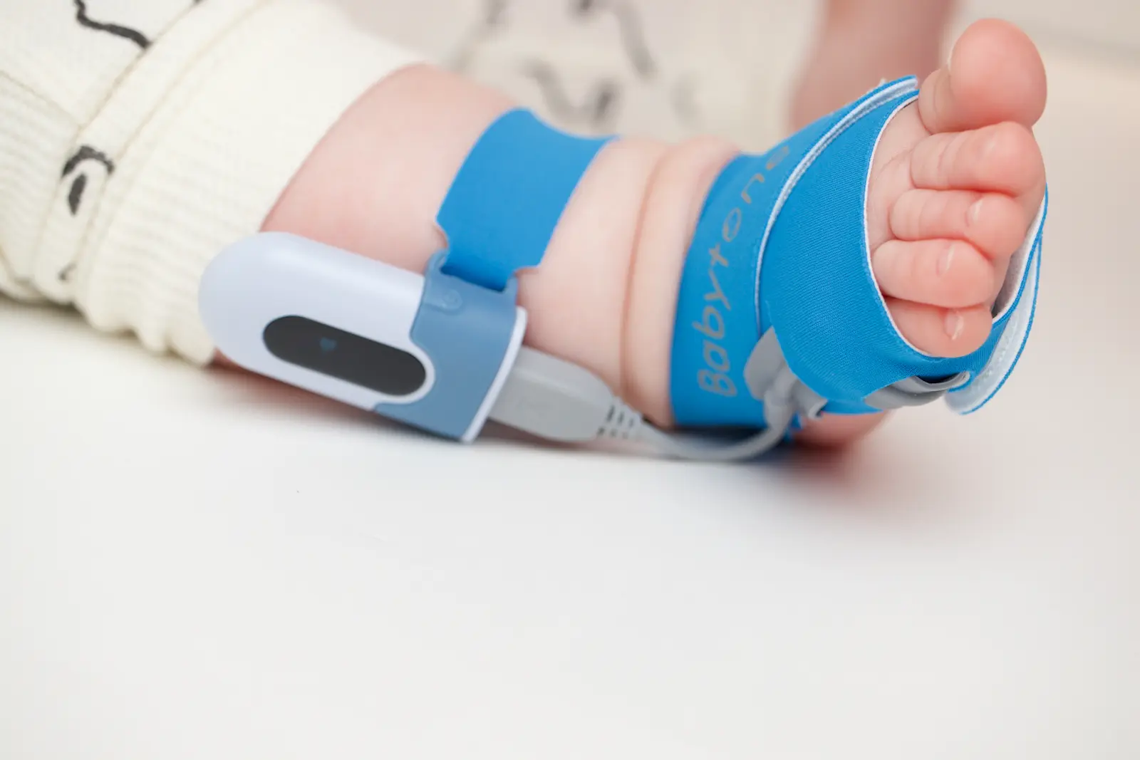 Baby foot wearing a Babytone pulse oximeter sensor secured with blue straps for infant oxygen monitoring