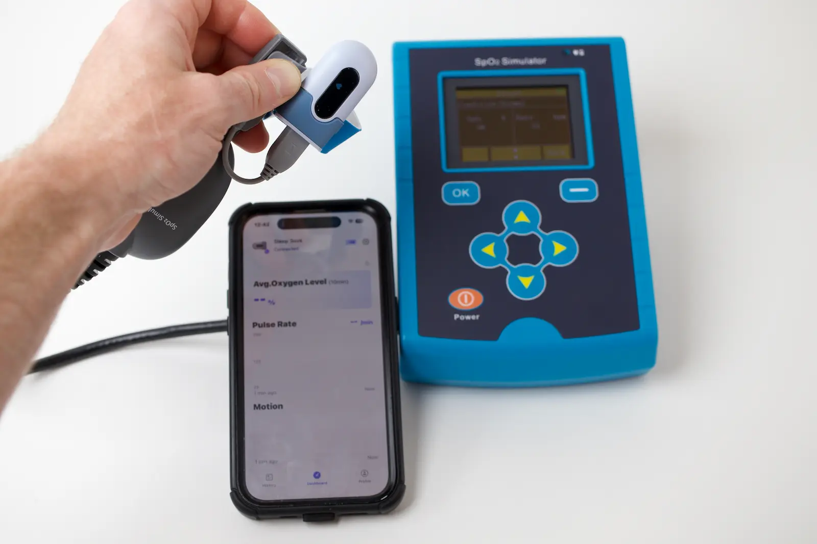 Hand holding pulse oximeter connected to SpO2 simulator beside smartphone showing oxygen level and pulse rate app